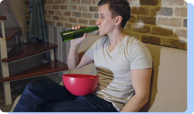 A man sitting on a couch in a dimly lit room, drinking from a green glass bottle (likely beer) with a red bowl of snacks in his lap.