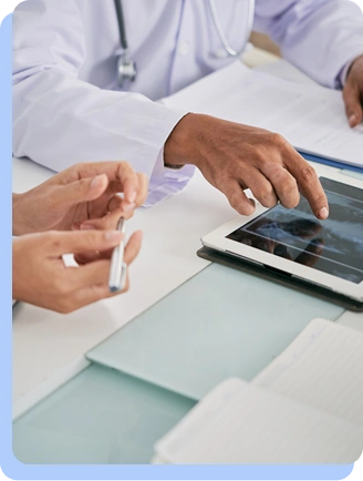 Close-up of a healthcare provider’s hands pointing to a digital tablet while consulting with a patient at a desk.
