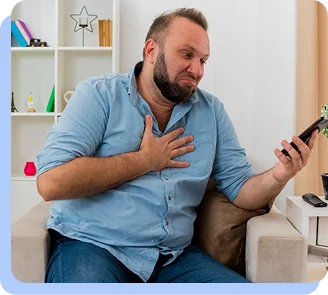 A middle-aged man sitting on a sofa looking distressed and grimacing in pain while clutching his chest with one hand and holding a smartphone in the other, suggesting an emergency health situation.