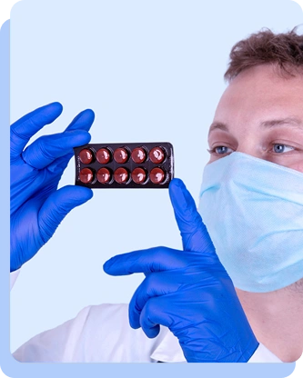 A person wearing blue medical gloves and a surgical mask, holding up a red blister pack of pills and pointing to it, suggesting a clinical or pharmaceutical context.