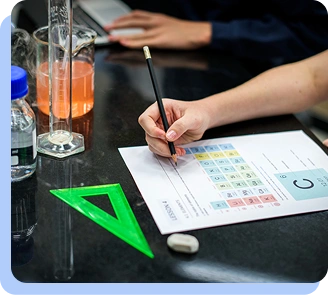 A close-up of a person's hand using a pencil to record data on a scientific chart or grid, with a green triangle ruler and laboratory glassware visible on the table.