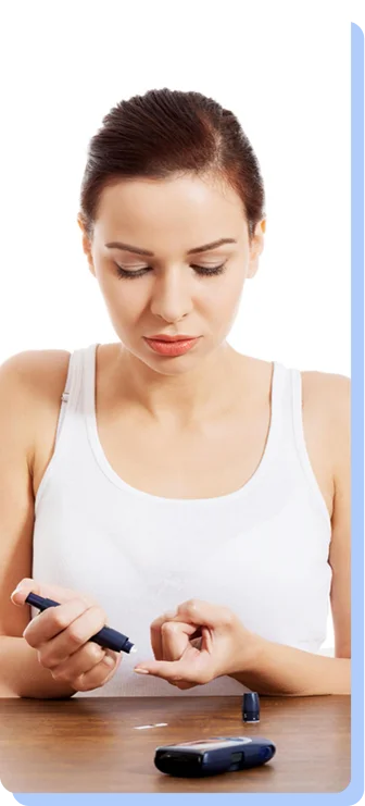 A woman in a white tank top sitting at a wooden table, focused on using a lancing device to draw a small drop of blood for a glucose test.