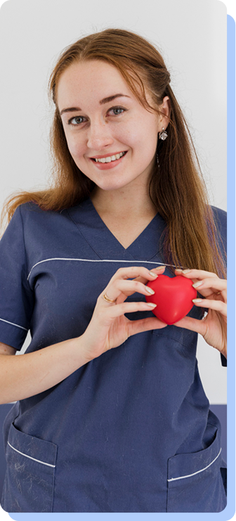 A young woman in blue medical scrubs smiling and holding a small red heart model with both hands near her chest.