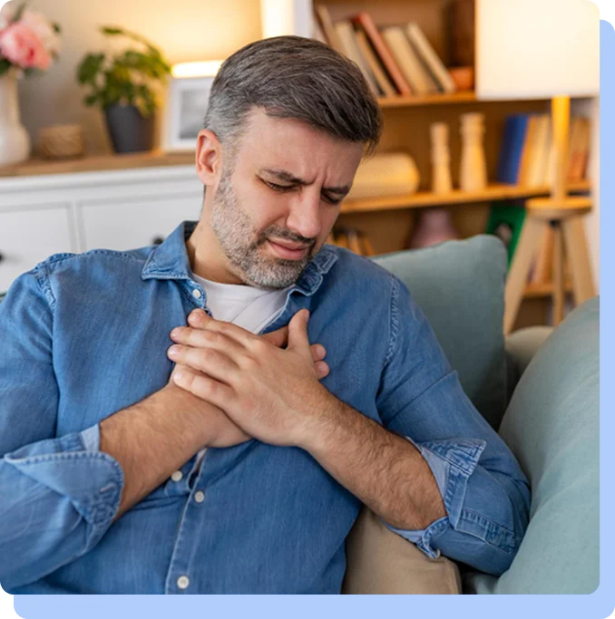 A middle-aged man sitting on a sofa, grimacing in pain and pressing both hands to his chest, depicting a symptomatic cardiac event in a home setting.