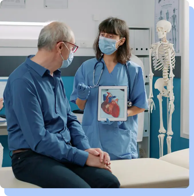 A female healthcare provider in blue scrubs and a face mask holding a digital tablet displaying an anatomical illustration of a human heart. She is showing the screen to an older male patient who is also wearing a face mask. A human skeleton model is visible in the background.