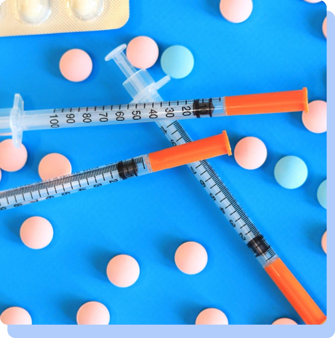 A flat-lay view against a vibrant blue background showing two medical syringes with orange caps crossing each other, surrounded by scattered pink and light blue round pills.