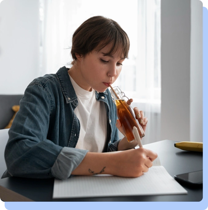 A person sitting at a desk with a notebook, drinking a brown beverage through a straw while writing, illustrating daily habits or "food noise."