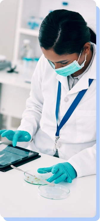 A scientist in a lab coat and green gloves works with a petri dish and a tablet in a laboratory setting.