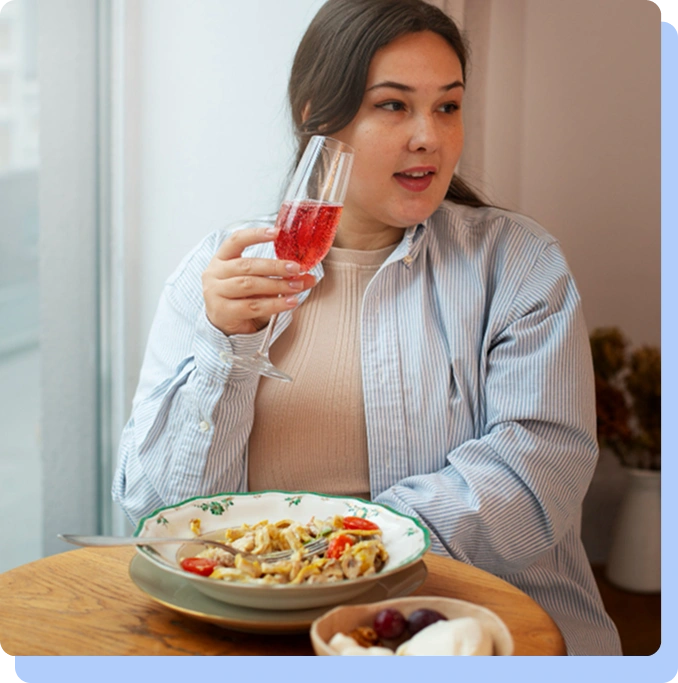 A woman sits at a wooden dining table, holding a glass of red sparkling juice. In front of her is a plate of pasta and a small bowl of fruit. She appears to be looking away from the food, illustrating the concept of reduced "food noise."