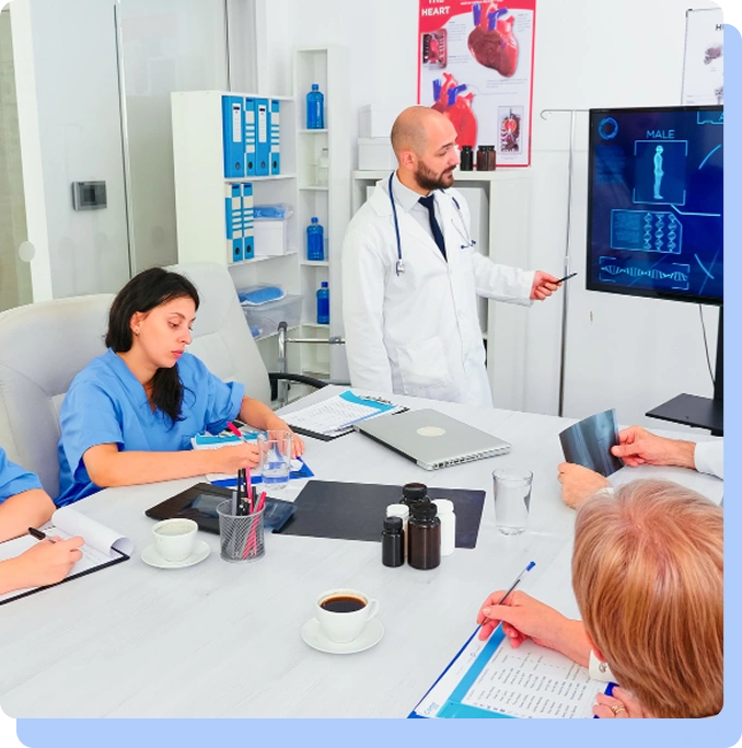 A group of medical professionals in blue scrubs and white lab coats are gathered around a conference table in a meeting room. A male doctor stands at the head of the table, pointing to a large digital screen displaying human anatomical data and heart diagrams.
