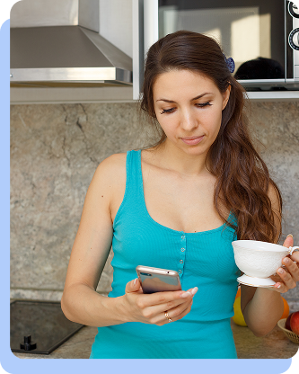 A woman in a blue tank top looking at her smartphone while holding a white teacup in a kitchen setting.