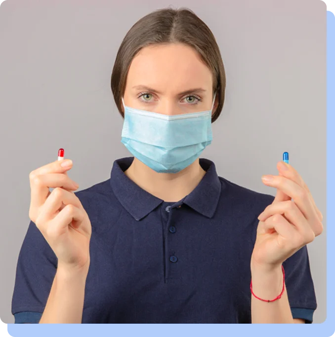A woman wearing a blue medical face mask and a dark polo shirt, holding a red and white capsule in one hand and a blue capsule in the other.