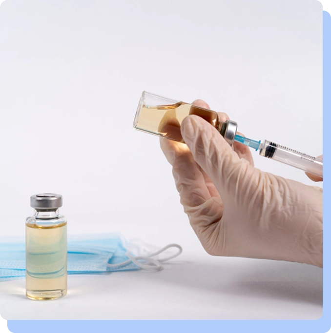 A close-up of a person wearing white medical gloves using a syringe to draw yellowish liquid from a small glass vial. Another identical vial and a blue surgical mask are visible in the blurred background.