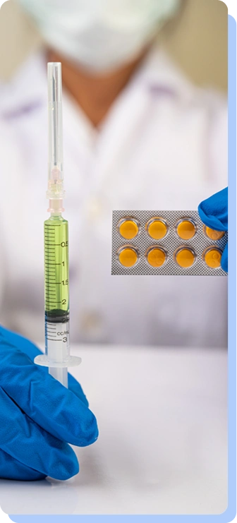 A person wearing blue medical gloves and a white lab coat holding a syringe filled with a yellowish-green liquid next to a gold blister pack of pills.