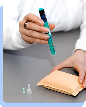 A close-up of a person's hands using a blue and green injection pen to practice an injection on a beige medical training pad.
