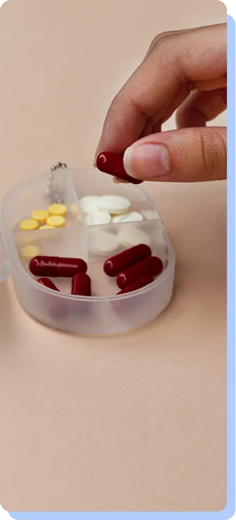 A hand picking up a large red capsule from a clear, circular multi-compartment pill organizer.