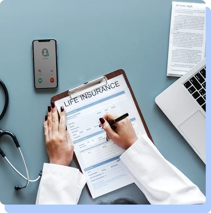 An overhead view of a doctor’s desk featuring a laptop, a smartphone, a stethoscope, and a hand filling out a "Life Insurance" application form.