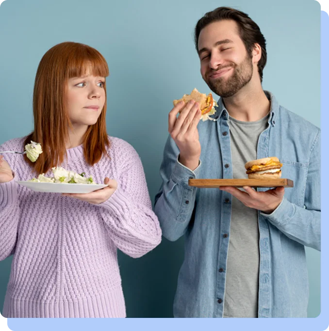 A man and a woman standing side-by-side against a blue background. The woman on the left looks skeptical while holding a plate of plain salad. The man on the right has his eyes closed in delight as he prepares to bite into a large breakfast sandwich, illustrating the difference between eating for fuel and eating for pleasure.