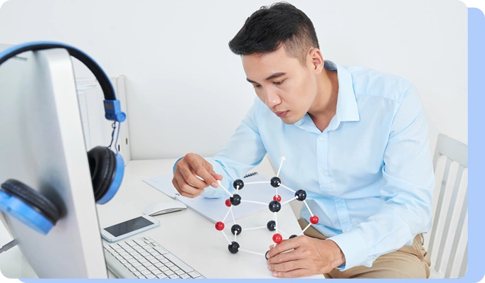 A young man in a light blue button-down shirt sitting at a desk, focused on assembling a complex 3D molecular ball-and-stick model. A computer and headphones are visible on his desk.