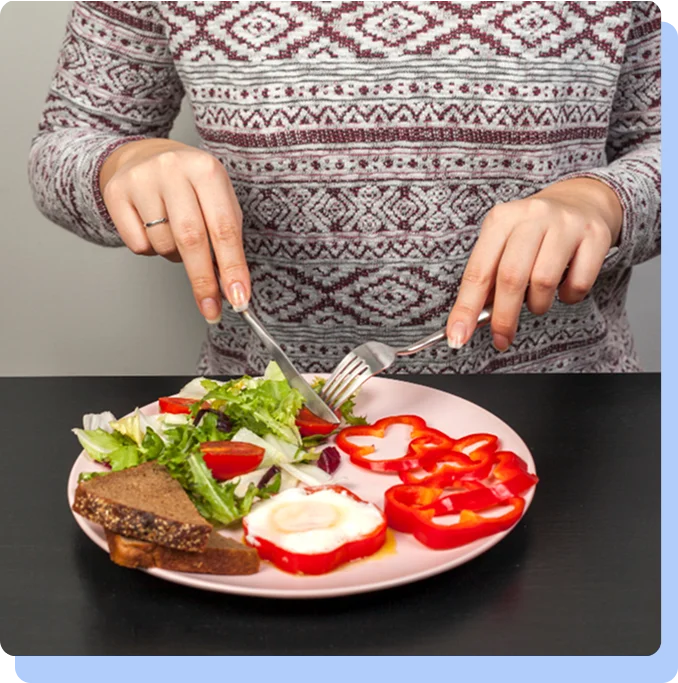 A person wearing a patterned sweater using a fork and knife to eat a healthy meal consisting of a fried egg inside a bell pepper ring, salad greens, and a slice of dark whole-grain bread.