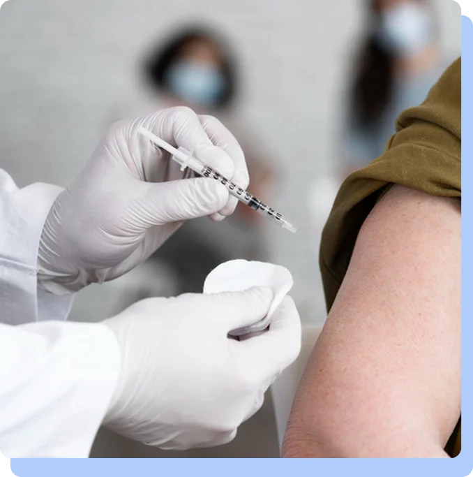 A close-up, focused shot of a healthcare provider’s hands in white gloves. They are holding a small syringe and a cotton swab, preparing to administer an injection into the upper arm of a patient. The background is softly blurred, showing a clinical setting.