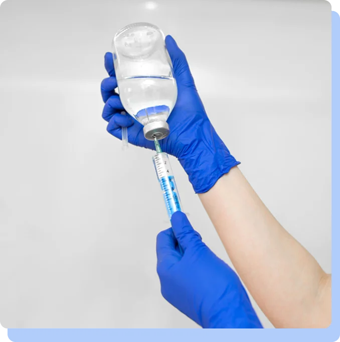 A close-up shot of hands wearing blue medical gloves. One hand holds a clear glass medication vial upside down while the other hand inserts a syringe needle into the rubber stopper to draw out liquid.