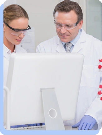 Two scientists, a man and a woman in white lab coats and safety glasses, looking intently at a computer monitor in a bright office or lab setting.