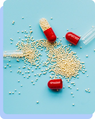 A top-down view of several red and white capsules spilled onto a light blue surface, with hundreds of tiny white medicinal granules scattered around them.