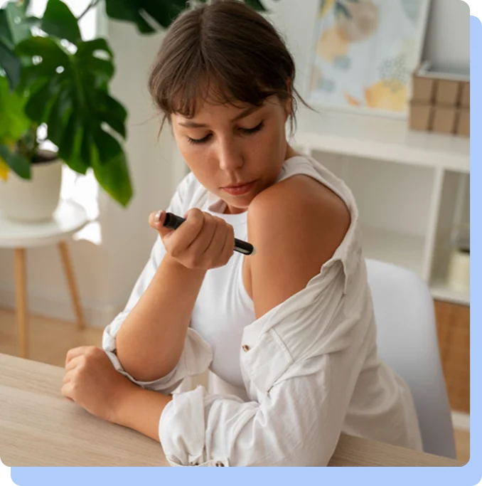 A woman in a white button-down shirt sitting at a desk, looking down at her arm as she prepares to administer an injection with a medication pen.