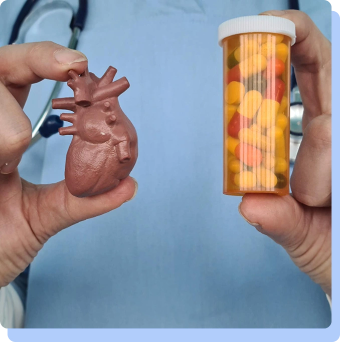 A close-up of a person's hands against a light blue medical scrub background. One hand holds a small, realistic model of a human heart, and the other holds a clear orange prescription pill bottle filled with various colorful capsules.