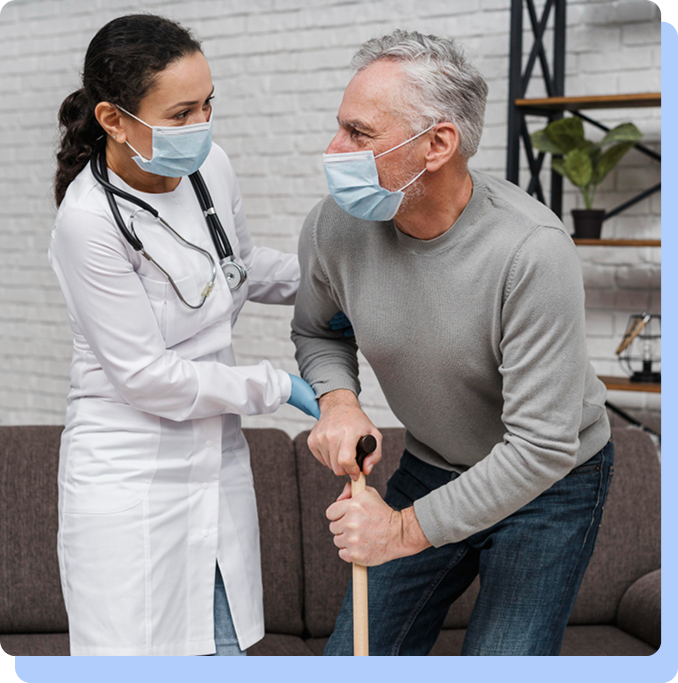 A female healthcare worker wearing a white coat and a surgical mask providing physical assistance to an elderly man. The man is also wearing a mask and using a wooden cane to stand up from a sofa.