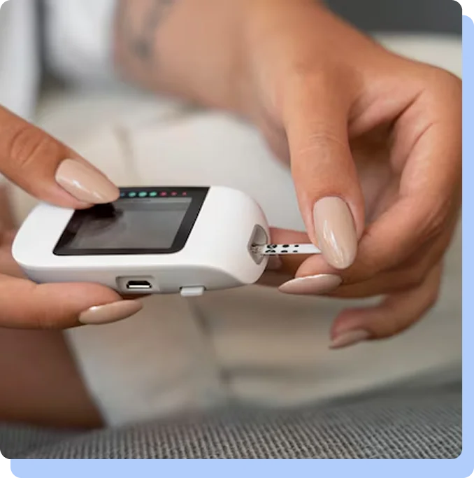 A close-up of a person's hands with manicured nails holding a white digital blood glucose meter. They are in the process of inserting or removing a test strip from the device.