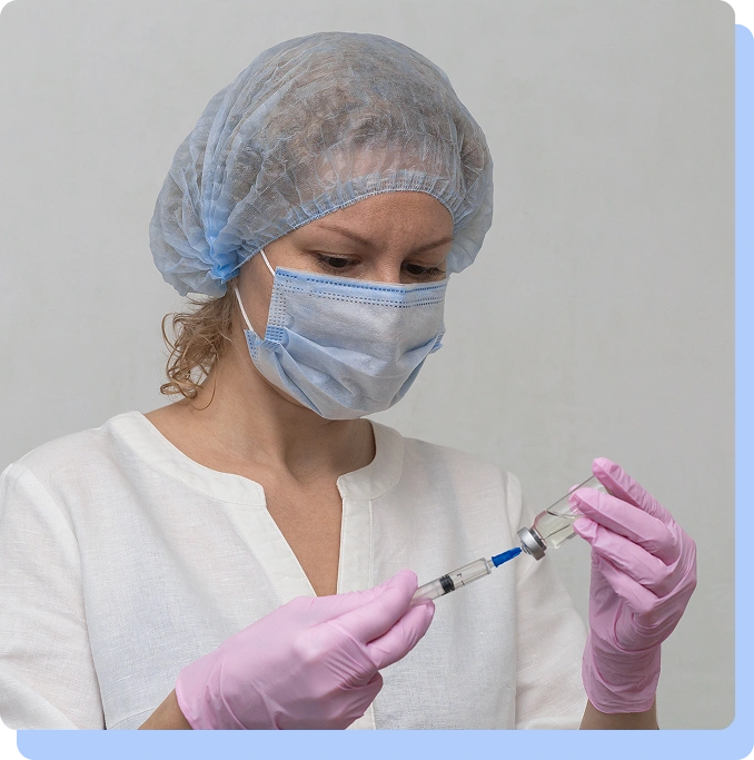 A healthcare worker wearing a surgical mask, hairnet, and pink gloves filling a syringe from a small vial.
