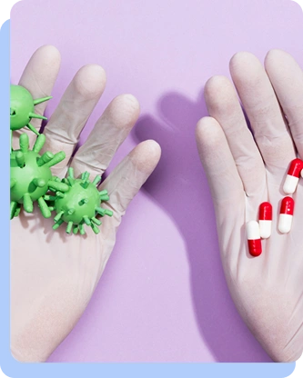 A pair of hands in white gloves shown against a purple background. The left palm holds green, spiked models representing virus or bacteria cells, while the right palm holds several red and white capsules.