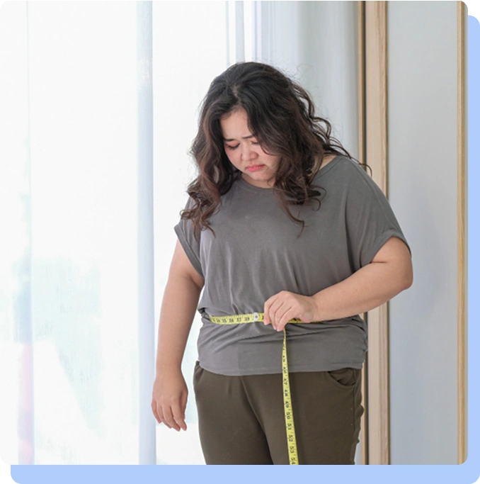 A woman standing in front of a bright window, looking down with a concerned expression as she uses a yellow measuring tape to check the circumference of her waist.
