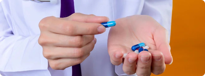 A close-up of a person in a white lab coat holding a single blue capsule in one hand while several other blue and white capsules sit in their open palm.