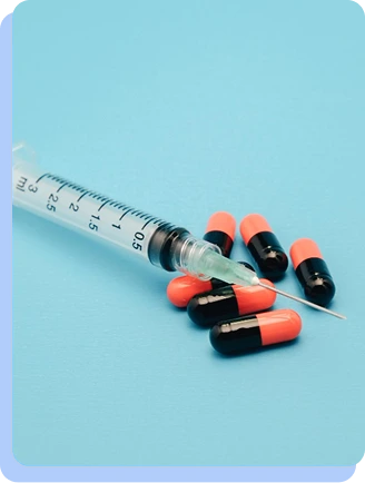 A medical syringe lying next to several orange and black capsules on a light blue background.