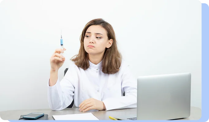 A woman in a white coat sitting at a desk with a laptop, holding a syringe up to the light with a skeptical or contemplative expression.