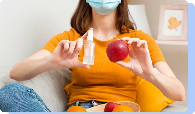 A person wearing a medical mask holding a small spray bottle and a red apple, with various fruits on the table in front of them.