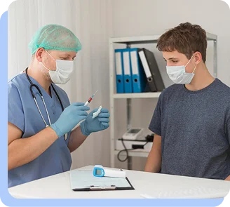 A male doctor in blue scrubs and a face mask speaking with a young male patient in a clinical office.