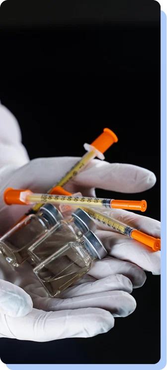 A close-up of a person’s cupped hands wearing white gloves, holding several small glass vials and orange-capped insulin syringes.