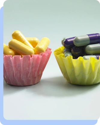 Two pink and green cupcake liners sitting side-by-side on a table, filled with yellow capsules and purple-and-grey capsules respectively.