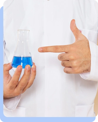 A person in a white lab coat holding a glass beaker containing a bright blue liquid in one hand while pointing at it with the other hand.
