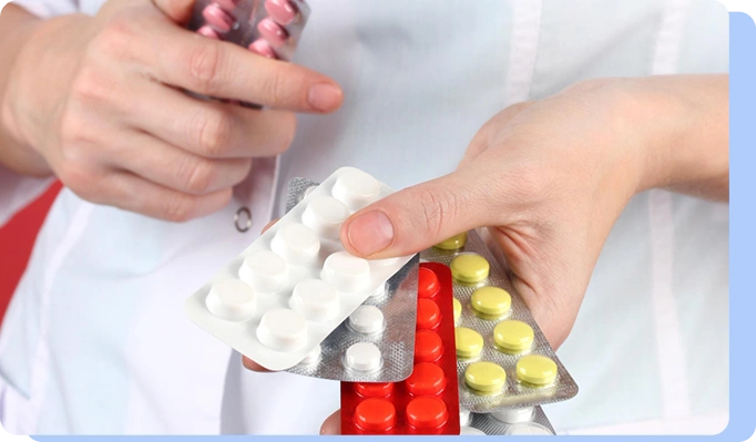 A person in a white lab coat holding multiple colorful blister packs of pills in various colors (white, yellow, and red).