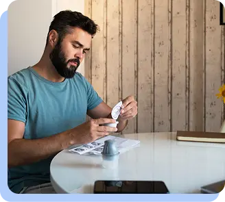 A man with a beard sitting at a white table, carefully examining a blister pack of medication and a small pill bottle.