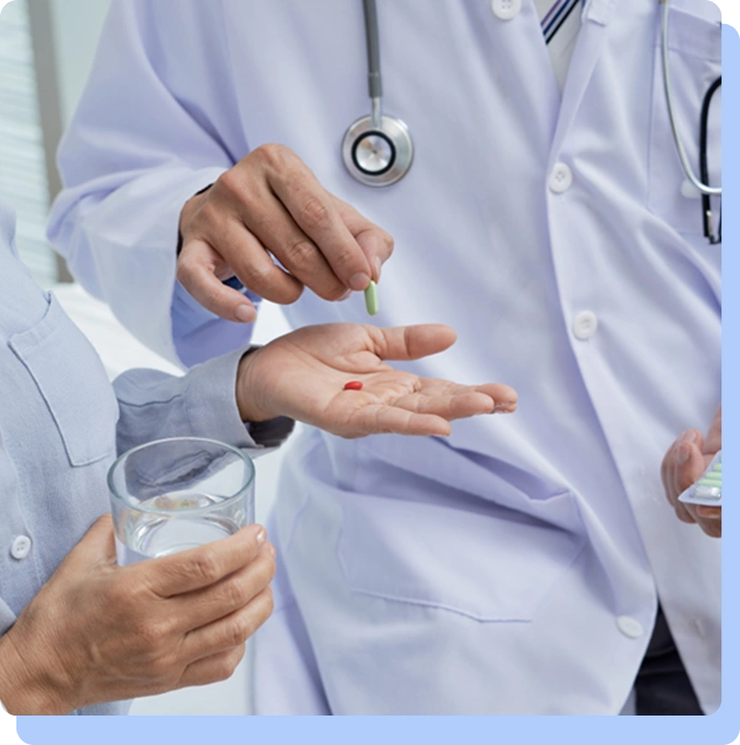 A close-up of a healthcare provider's hands holding a glass of water and offering two small red pills to a patient.