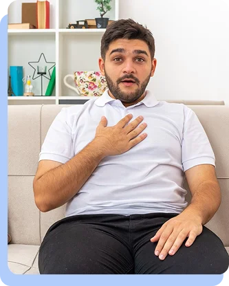 A man in a white polo shirt sitting on a couch, looking surprised or alarmed with one hand on his chest.
