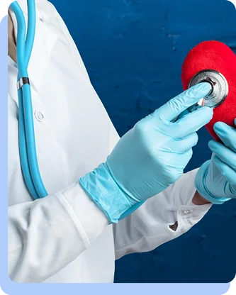 A healthcare professional in a white coat and blue gloves using a stethoscope to listen to a large, red heart-shaped model.