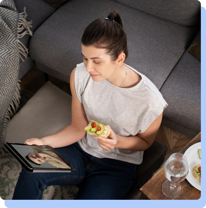 An overhead view of a woman sitting on the floor, holding a piece of avocado toast while looking at a tablet showing a person on a video call.