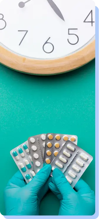 Hands in blue medical gloves holding several different blister packs of medication in front of a large wall clock.